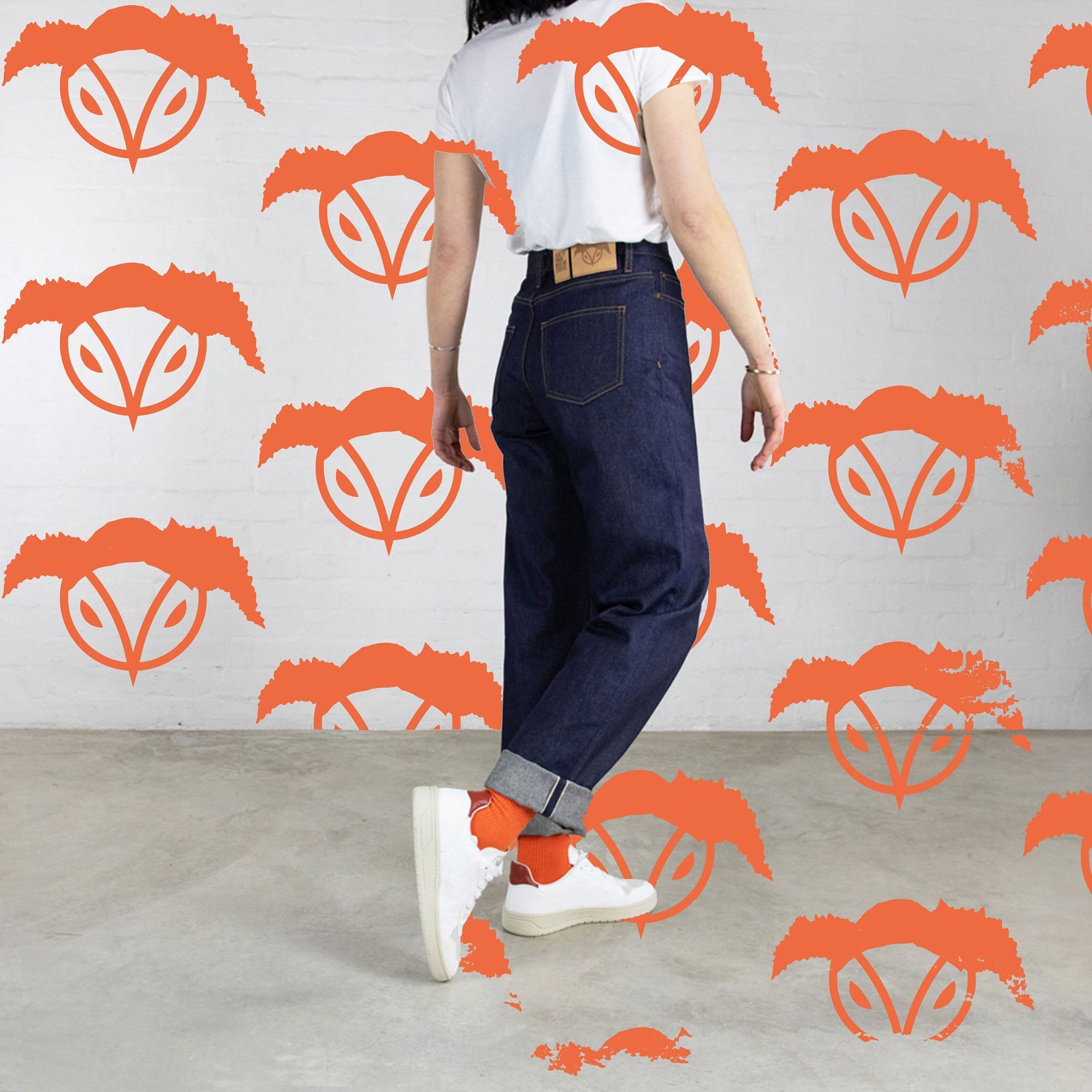 Person standing against a white wall with orange graphic logos, wearing a white T-shirt, dark blue jeans with rolled cuffs, orange socks, and white sneakers.