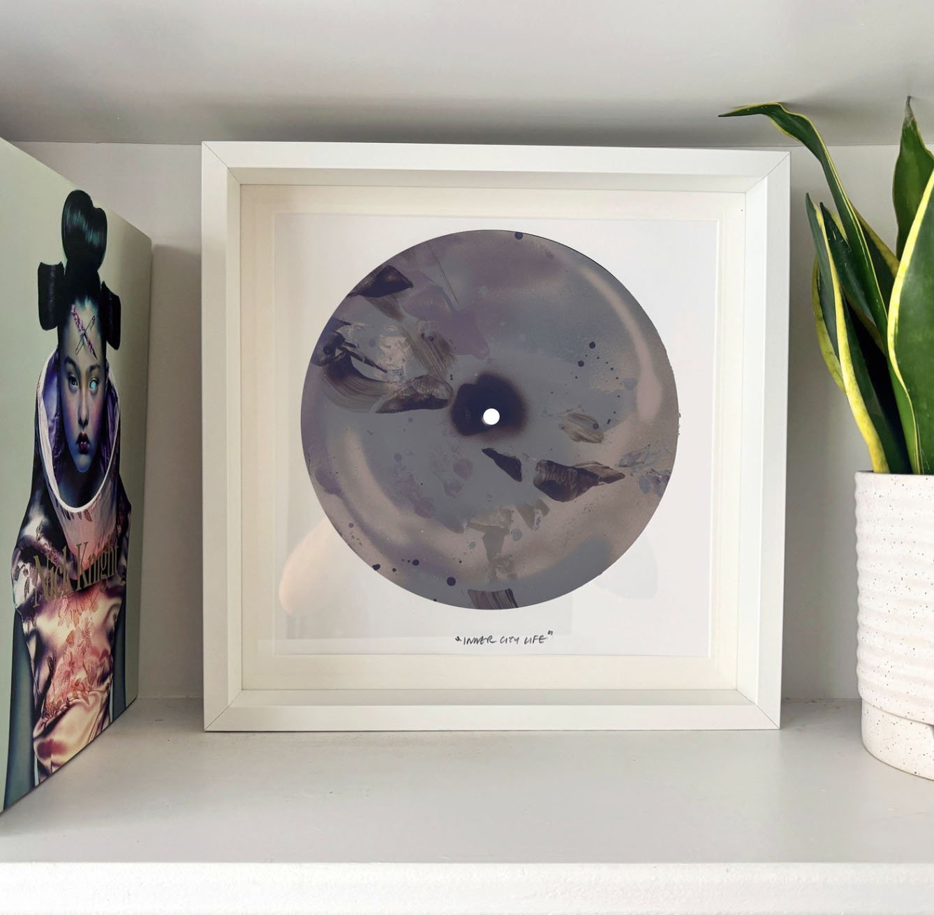 Decorative framed print titled "Inner City Life" with abstract circular design in shades of gray, black, and white, placed on a white shelf next to a colorful artwork of a woman with vibrant makeup and elaborate hairstyle, and a potted snake plant with green and yellow leaves.