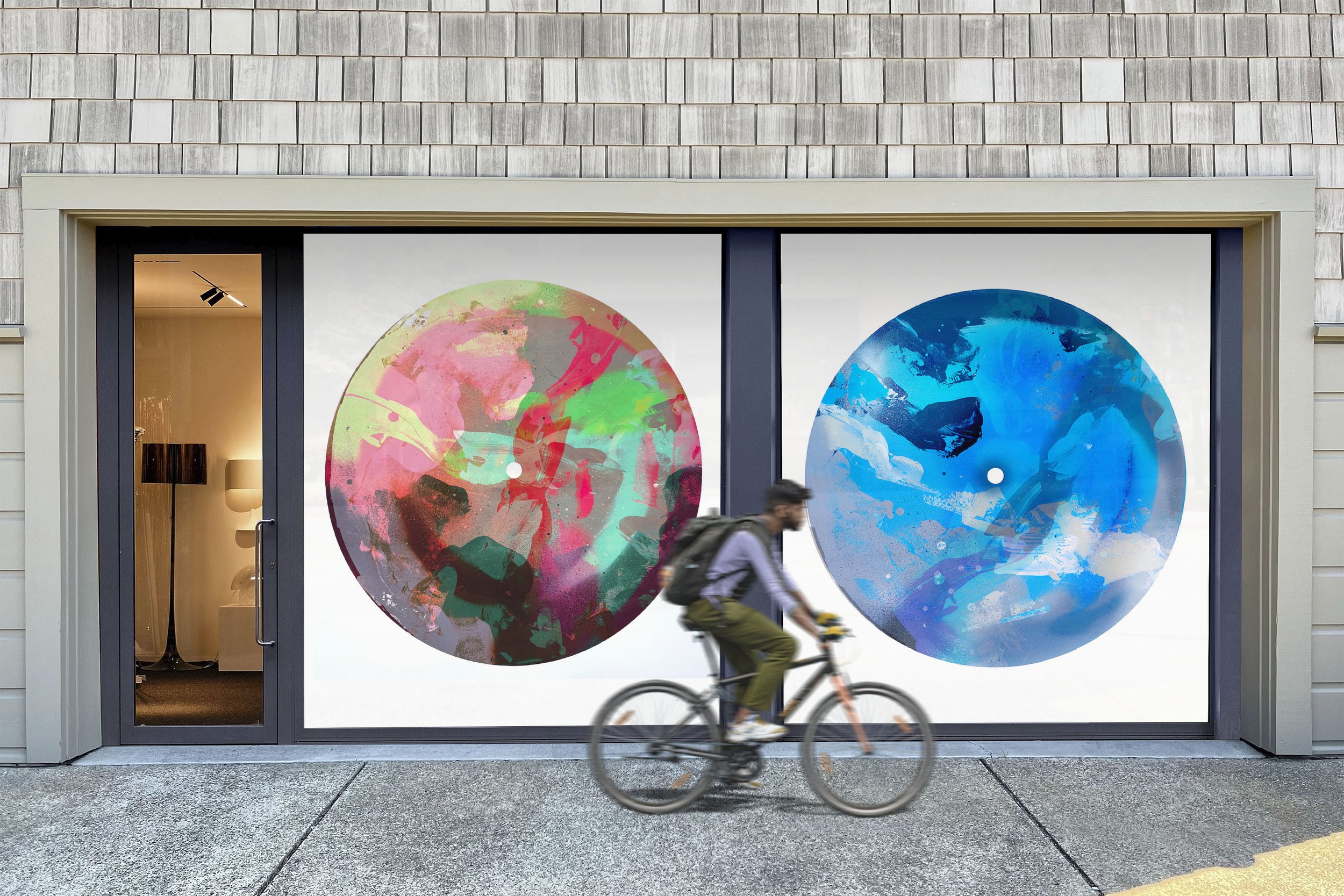 Storefront window with two large abstract art circles, a colorful pink and green one on the left and a blue one on the right, with a cyclist riding by in front of the window.