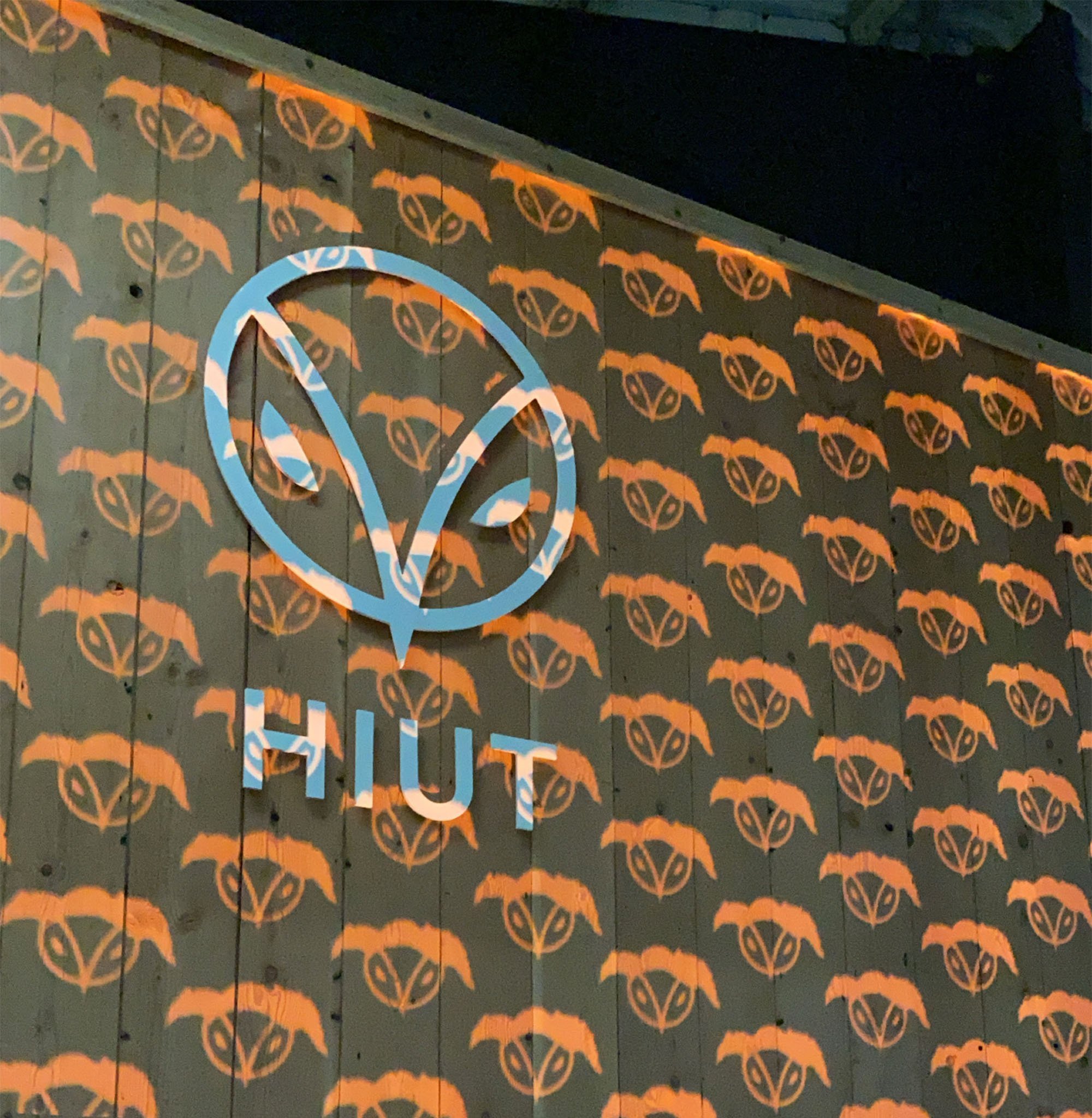 A wall decorated with repeated orange logos of a bat and a skull pattern, with a large illuminated sign displaying a stylized 'V' and the word 'HUT' underneath.