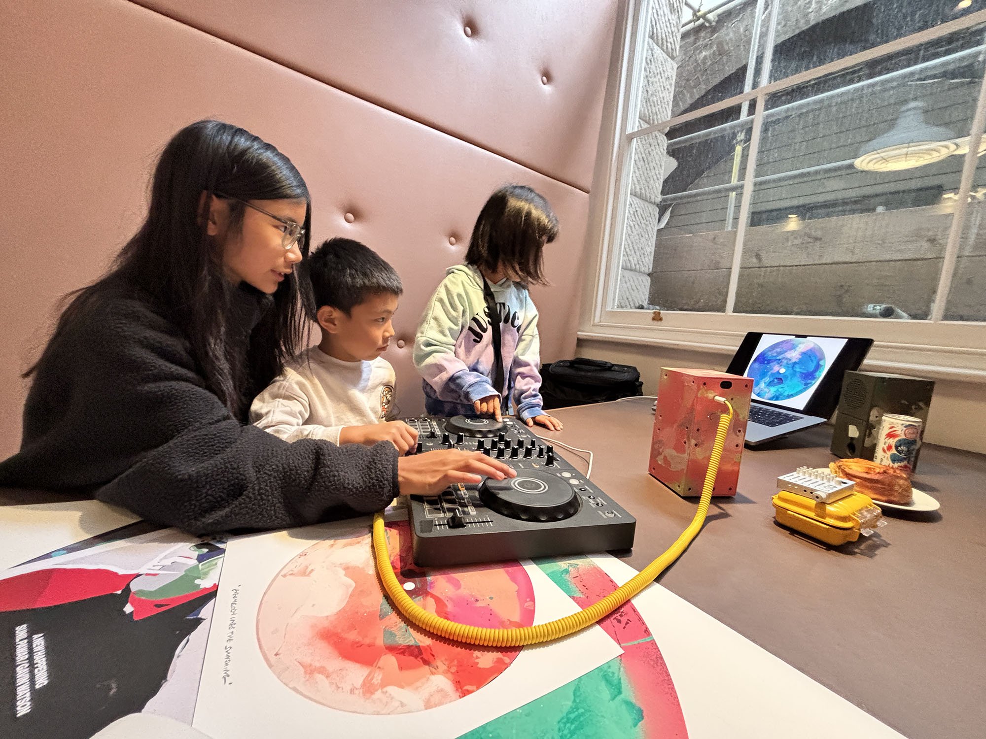 A woman and two children sitting at a table with a DJ controller and laptop, looking at the screen. The woman is wearing glasses and a black sweater, and the children are focused on the DJ equipment. There are snacks and colorful items on the table, with a large window in the background showing outside.
