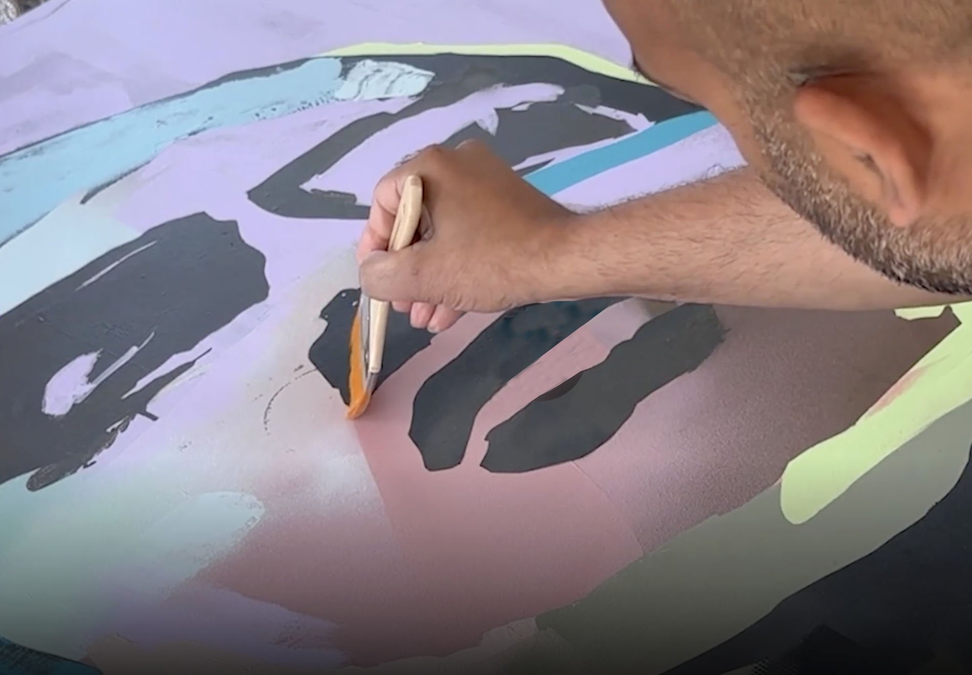 An artist is painting an abstract colorful artwork on a canvas. The artist is using brushes to add details to the painting, which features various shapes and colors, including black, pink, yellow, and blue.