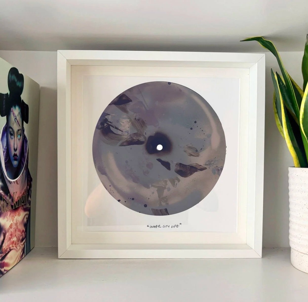 Framed abstract artwork titled 'Inner City Life' featuring gray and black swirling patterns and paint splatters on white background, surrounded by plant and poster.