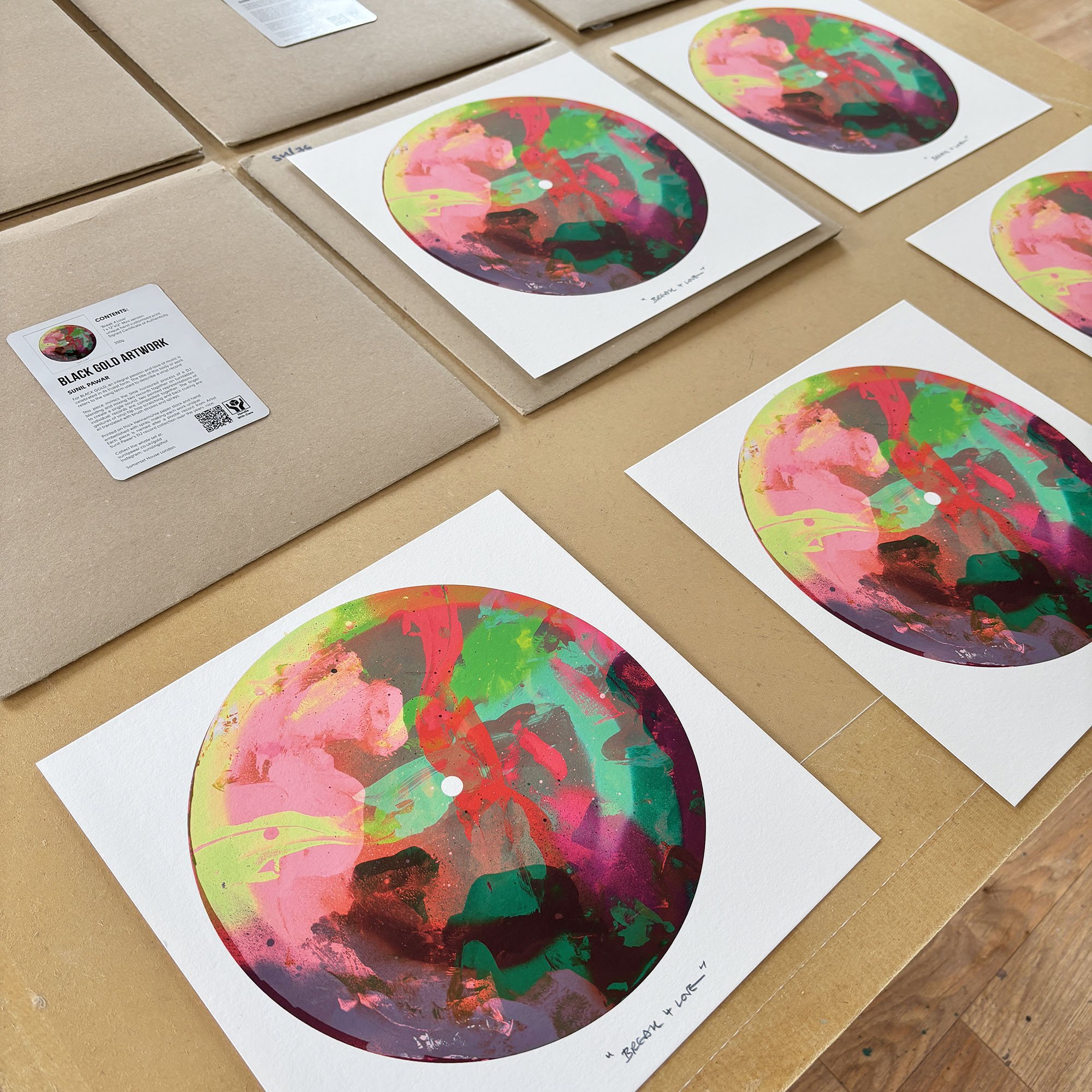 Multiple prints of abstract, colorful artwork with splashes of pink, green, red, and black, laid out on a table along with an art packaging label.