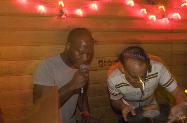 Two people singing karaoke in a dimly lit room with wooden walls and red lights.