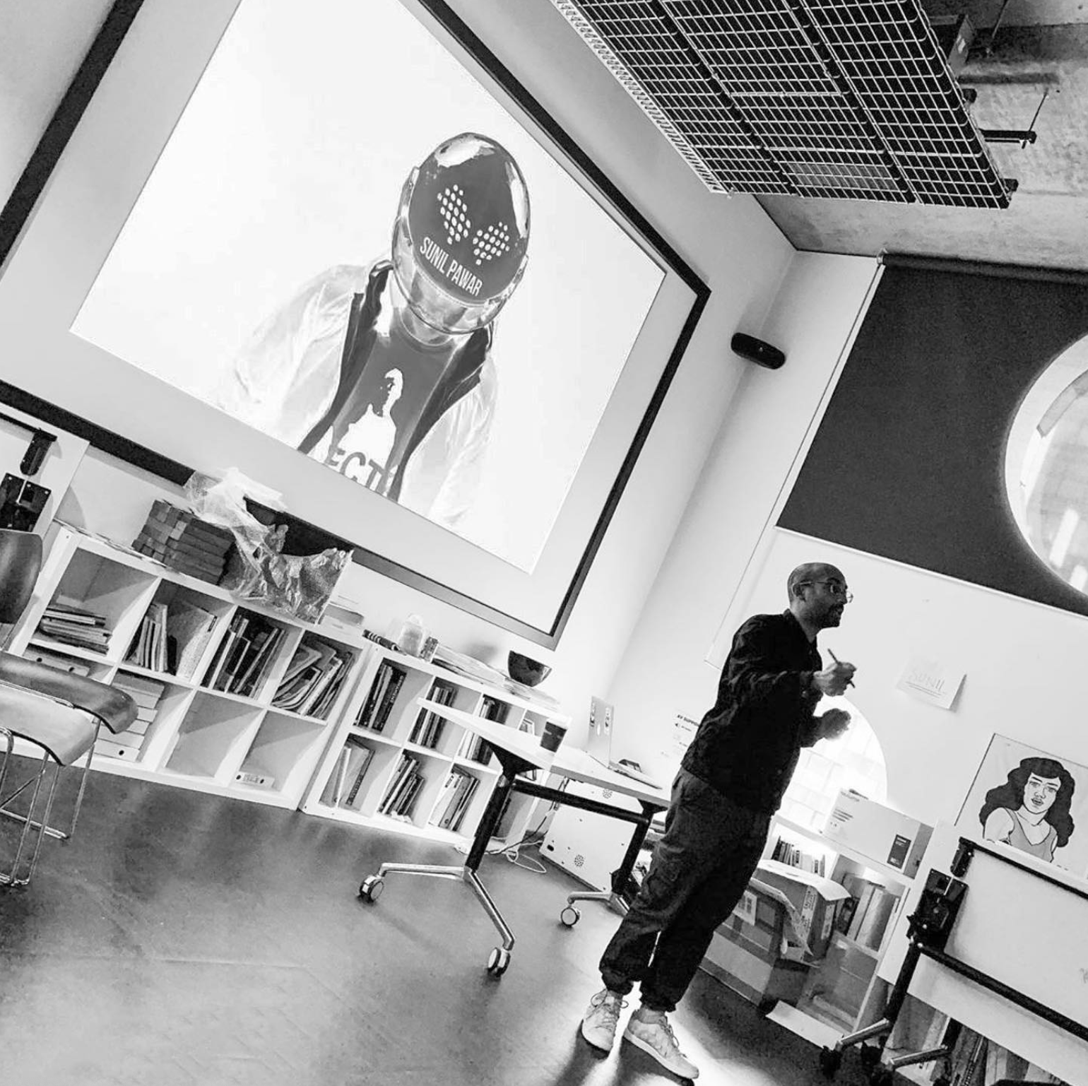 A man standing in a room with bookshelves and artwork, holding a drink and a straw, with a large digital screen displaying a person wearing a helmet behind him.