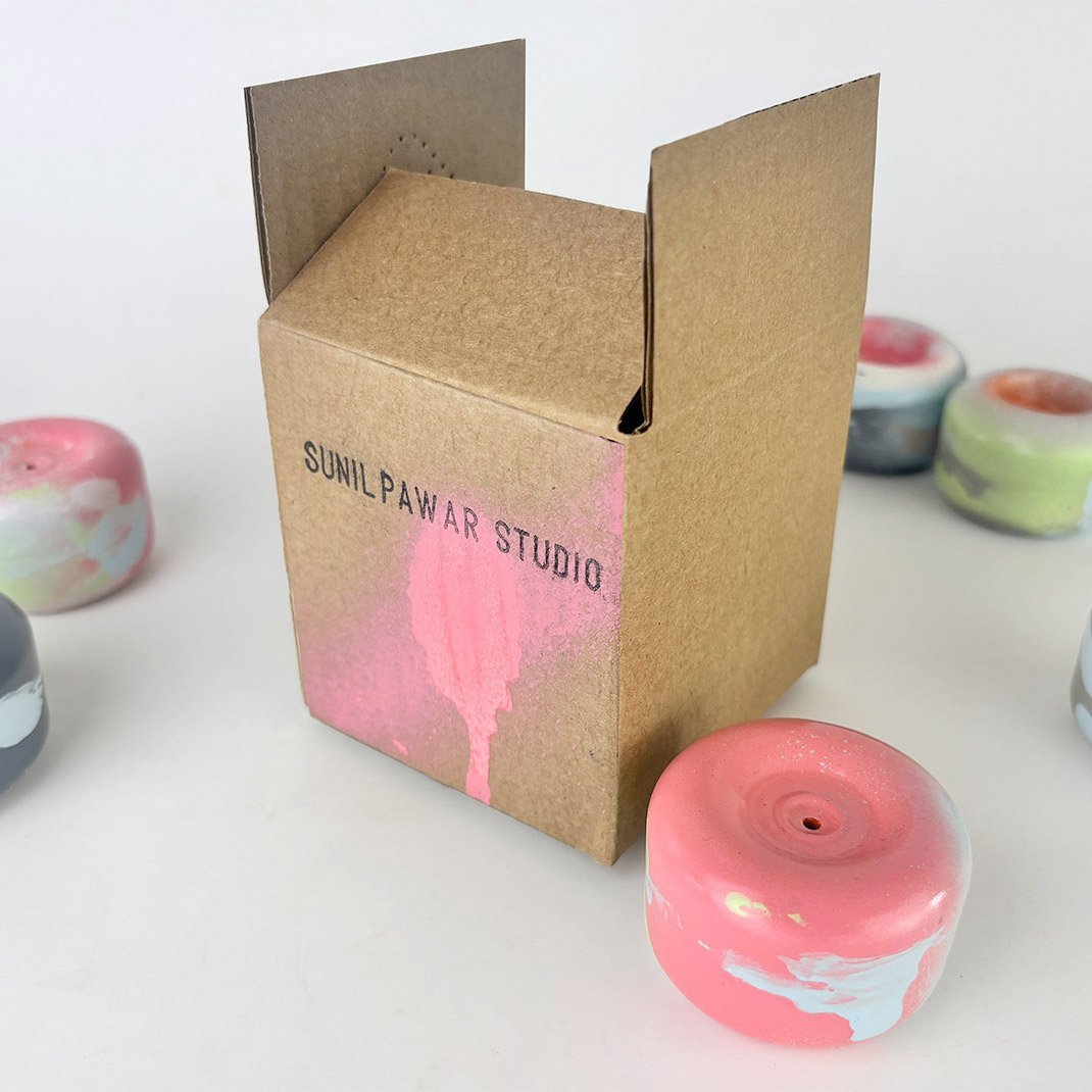 A cardboard box with a pink paint stain and the text 'SUNILPAWAR STUDIO' printed on it, surrounded by marbled pastel-colored candles on a white surface.