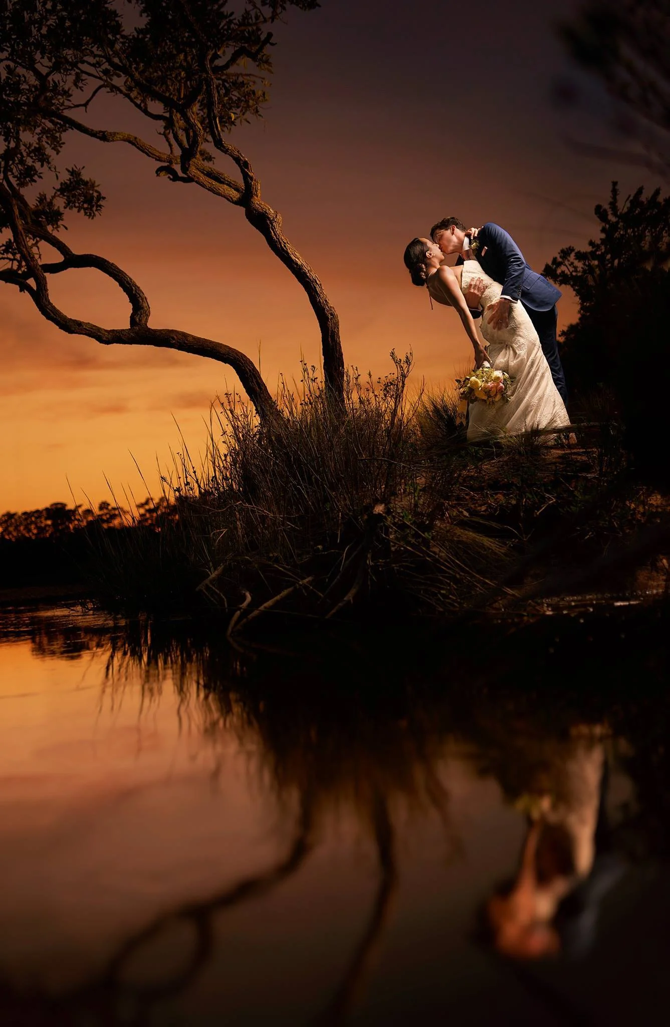 I love this shot mostly because it was damn tricky to get in position to shoot it. I had to lie on my stomach in an ant bed (in my defense I didn't realize it was there until about 45 seconds later!) and reach out over water. I've actually included a photo of me shooting it in the comments, my assistant was being sneaky. The symmetry of the bride and groom leaning over and the tree bent out over the water. I'd love to say that I planned that, but he grabbed her and went in for the dip without me even suggesting it! - Nicholas Gore