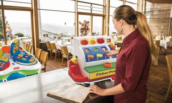 Eine Kellnerin in einem Restaurant bedient eine Fisher-Price-Spielzeugkasse. Satirisches Bild aus Das Letzte Wiki.