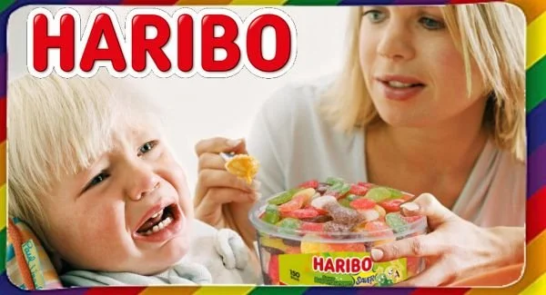 Parodie-Meme saurer Haribo-Süßigkeiten, die einem unbegeistert dreinblickenden Kind von seiner Mutter gefüttert werden. Satirische Fotomontage von "Das Letzte Wiki".