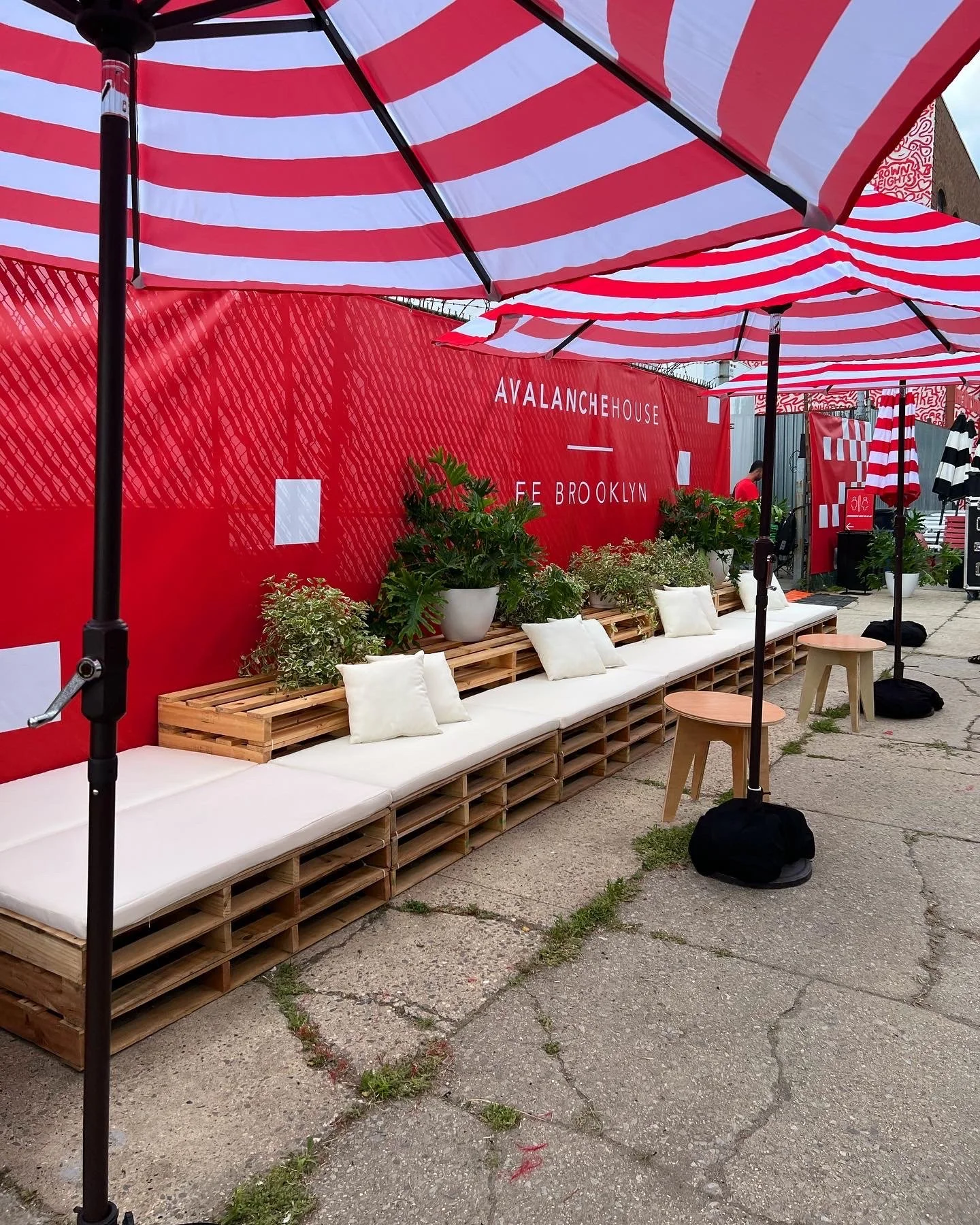 A long continuous sideline view pallet lounge with cream cushions and plants for an event in Brooklyn. Other rental furniture complimenting the lounge are side tables and umbrellas