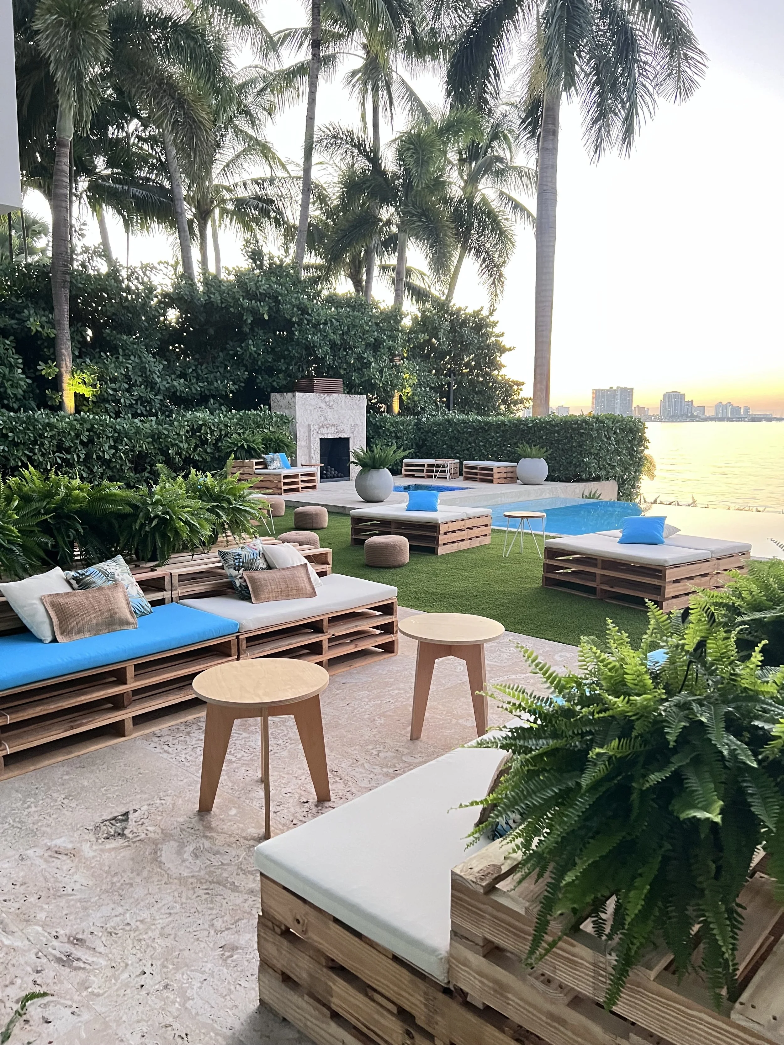 Pallet Sofas for rent set up with colorful cushions, tables, and plants, next to a pool with the Miami skyline in the background