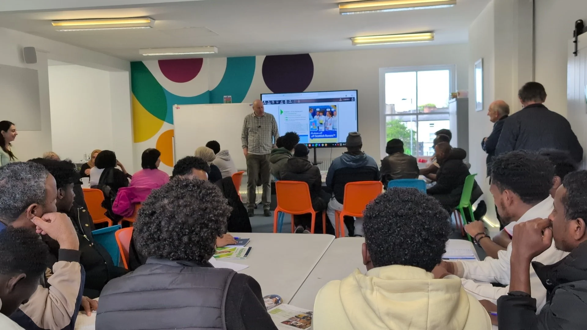 A group of people looks towards a teacher in a classroom. It is brightly decorated and there is a large TV screen they are looking at.