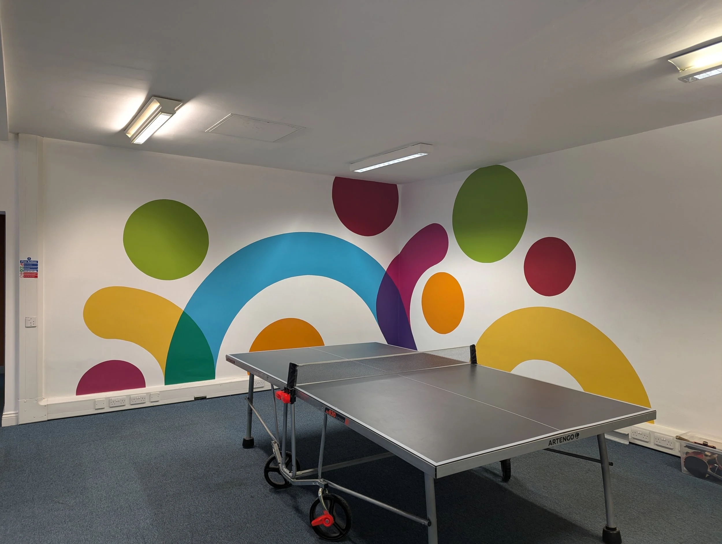 A photo of a table tennis table in front of a wall that is painted with shapes in all different colours
