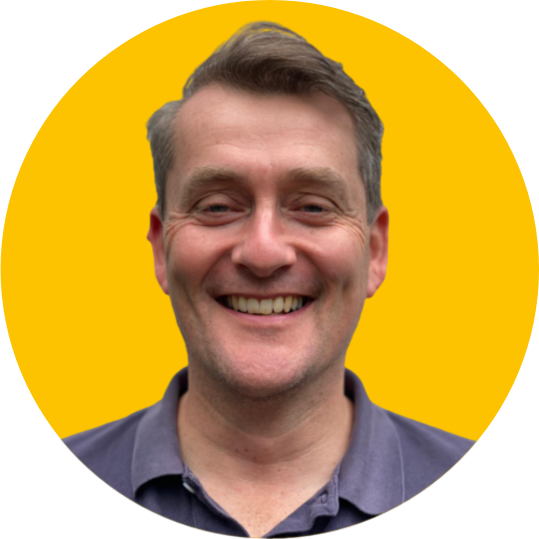 A man with grey hair smiles at the camera. He is wearing a grey polo shirt and is set against a yellow background.