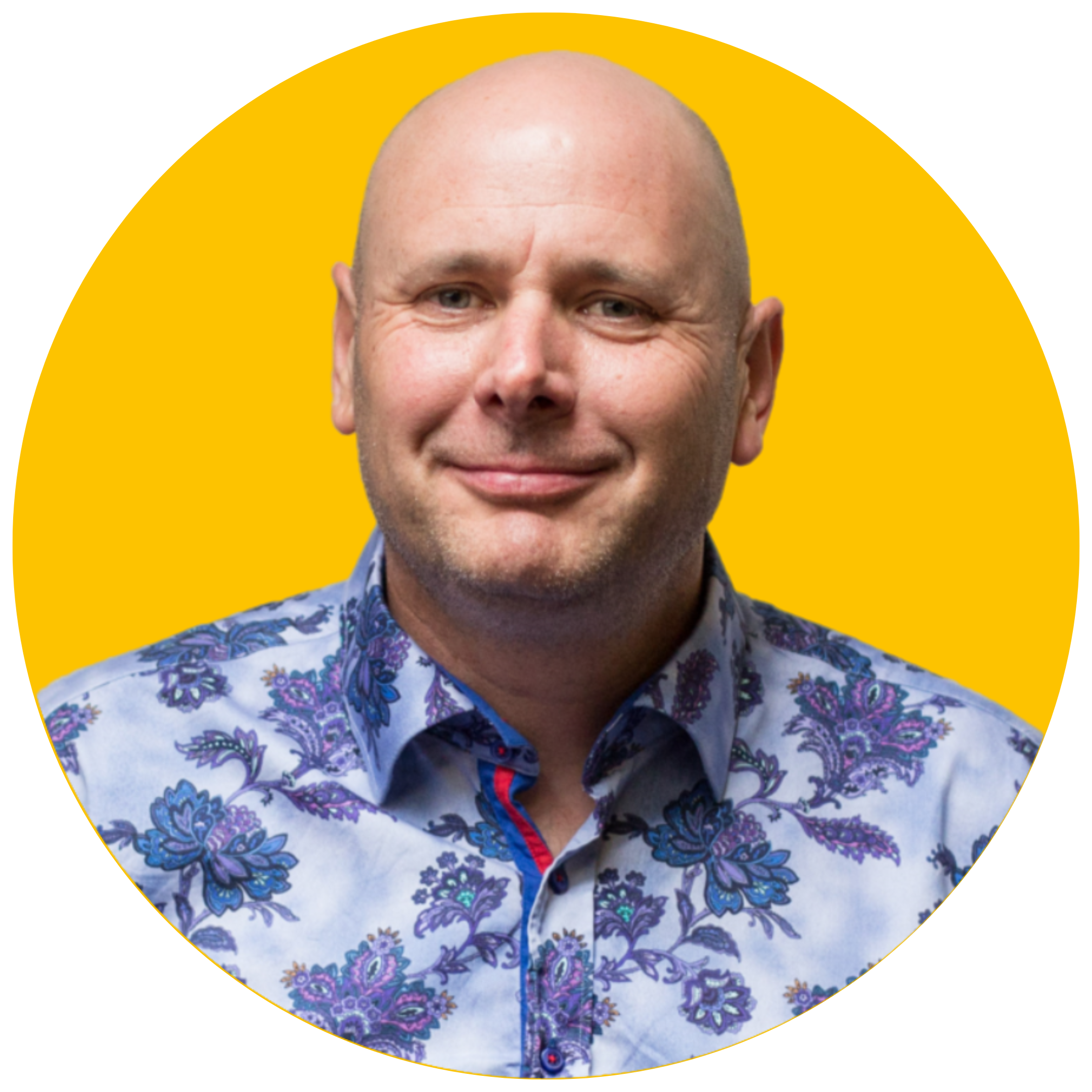 A bald man smiles at the camera. He is wearing a blue floral shirt and is set against a yellow background.