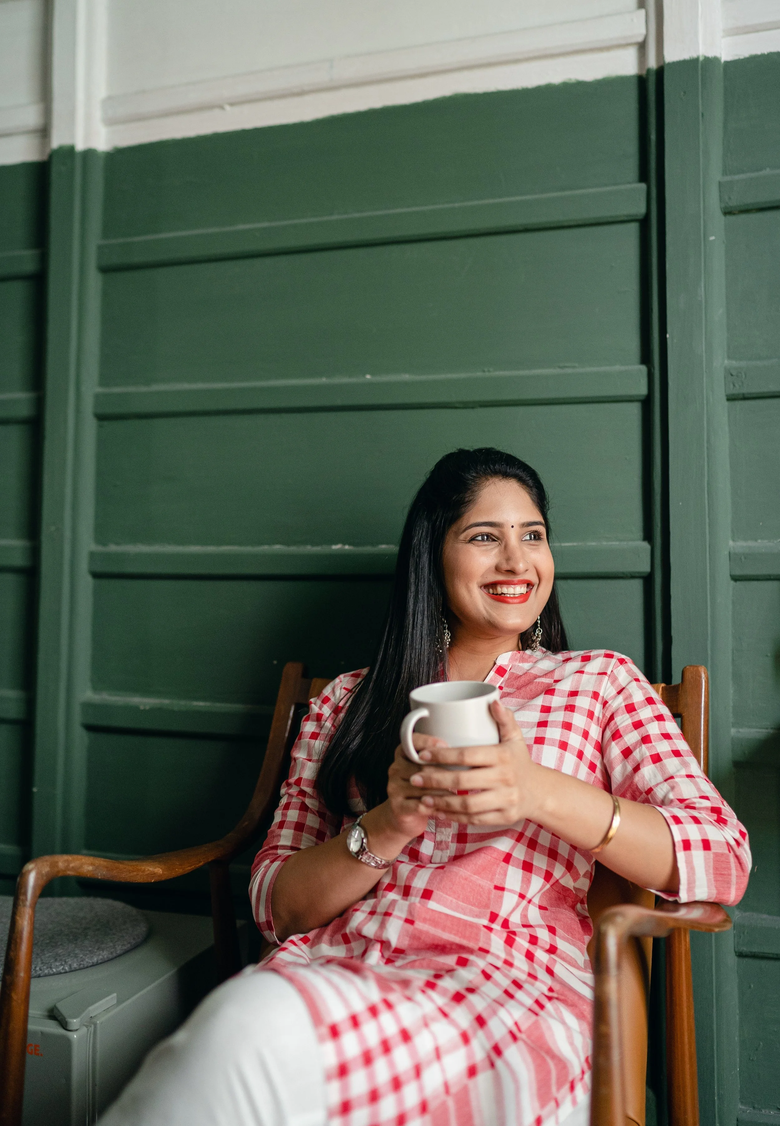A woman with long dark hair smiles in front of a green wall. She is holding a mug and wears a red checked shirt.