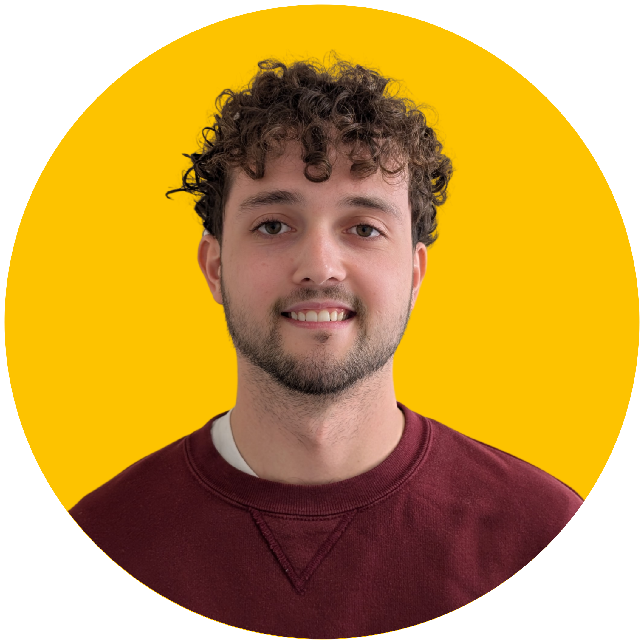 A man with curly brown hair smiles at the camera. He is wearing a red jumper, and is set against a yellow background.