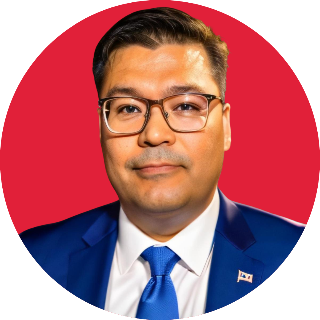 A man with short dark hair and glasses smiles at the camera. He is wearing a blue suit with a white shirt and blue tie, and is set against a red background.