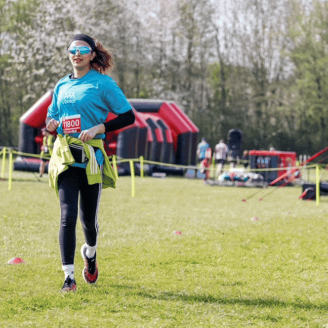 A woman wearing leggings and a blue T Shirt jogs on a green field with inflatable obstacles in the background.