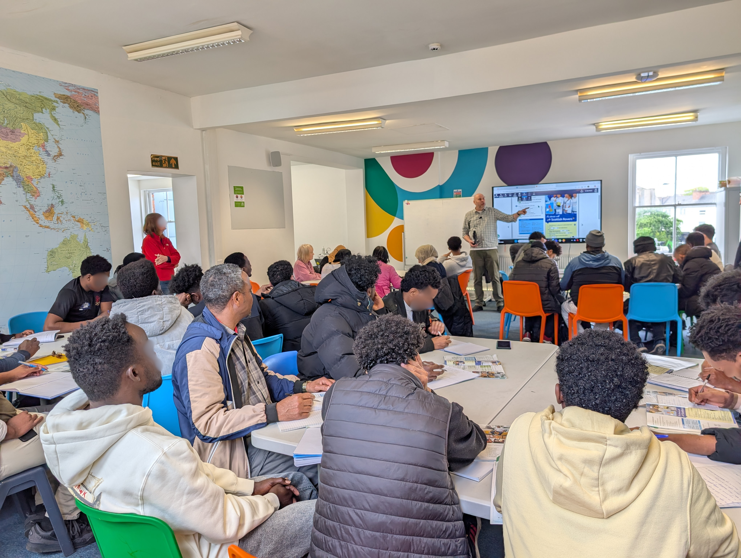 a photo of people sitting in a class learning English with a teacher standing at the front of the class