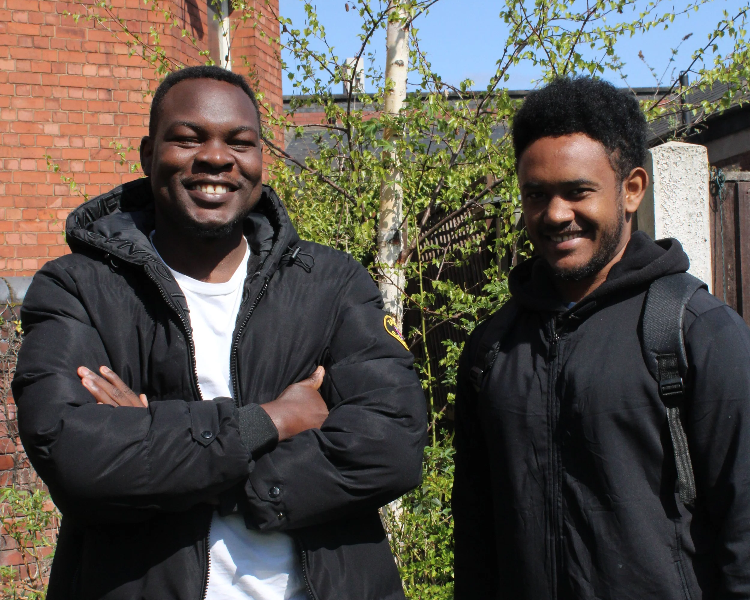 Two men stand in a garden smiling, in bright sunlight. They are wearing dark, winter clothing and surrounded by greenery.