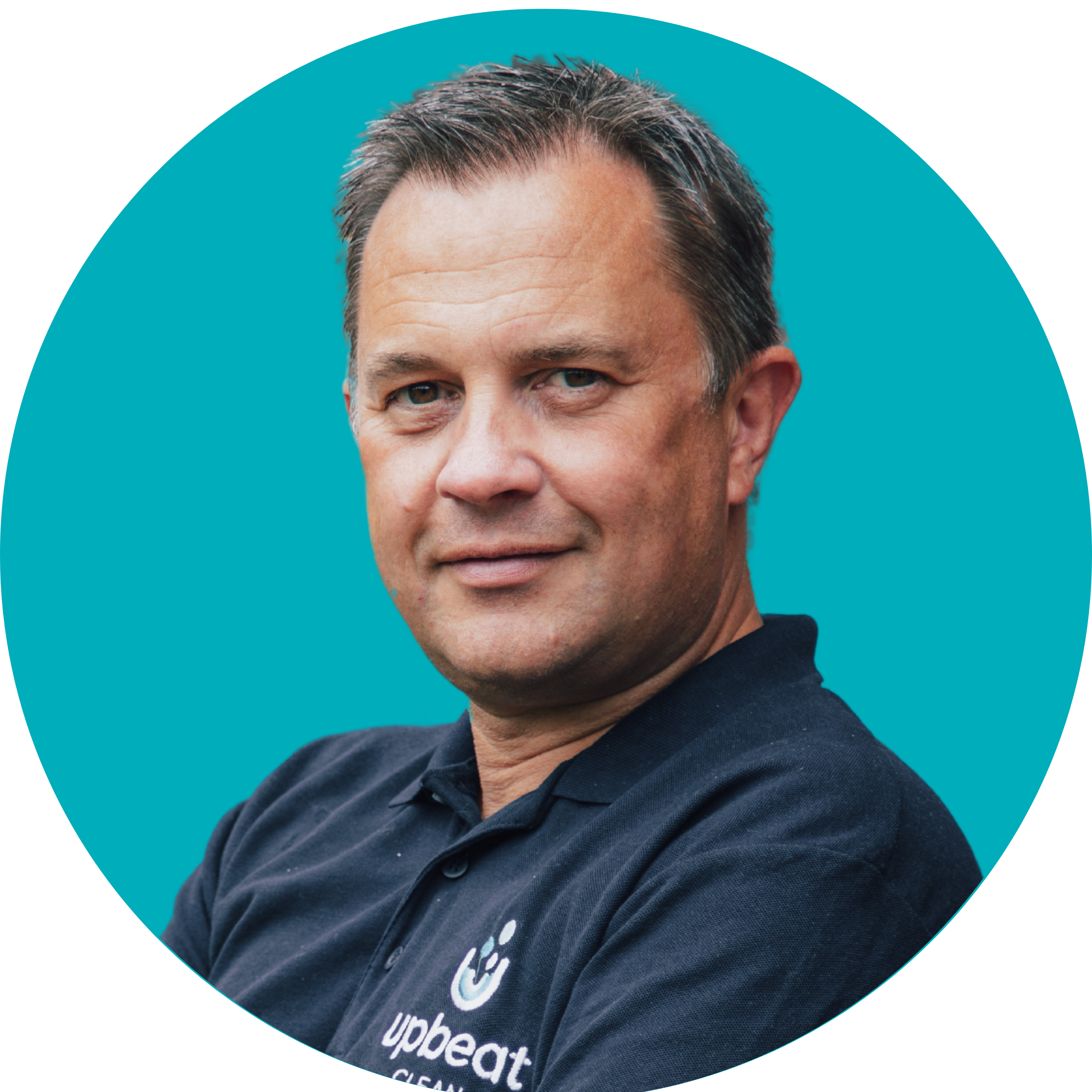A man with grey hair smiles at the camera. He is wearing a dark blue polo shirt, and is set against a teal background.