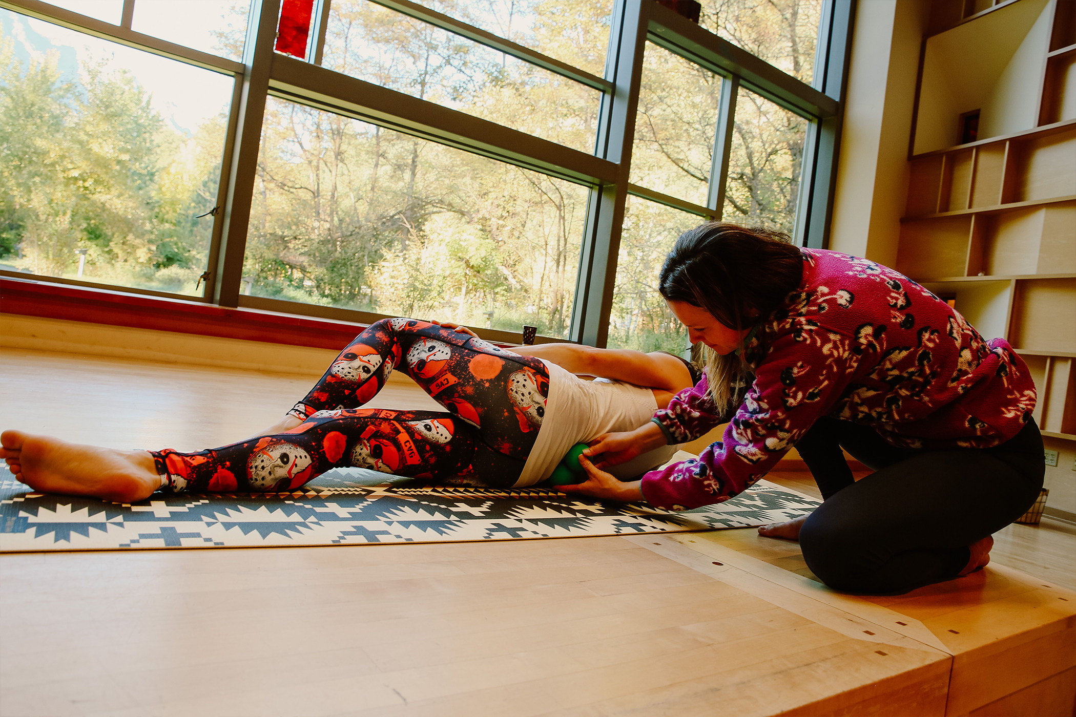A woman assisting a person lying on a mat with stretching or physical therapy in a room with large windows and a view of trees.