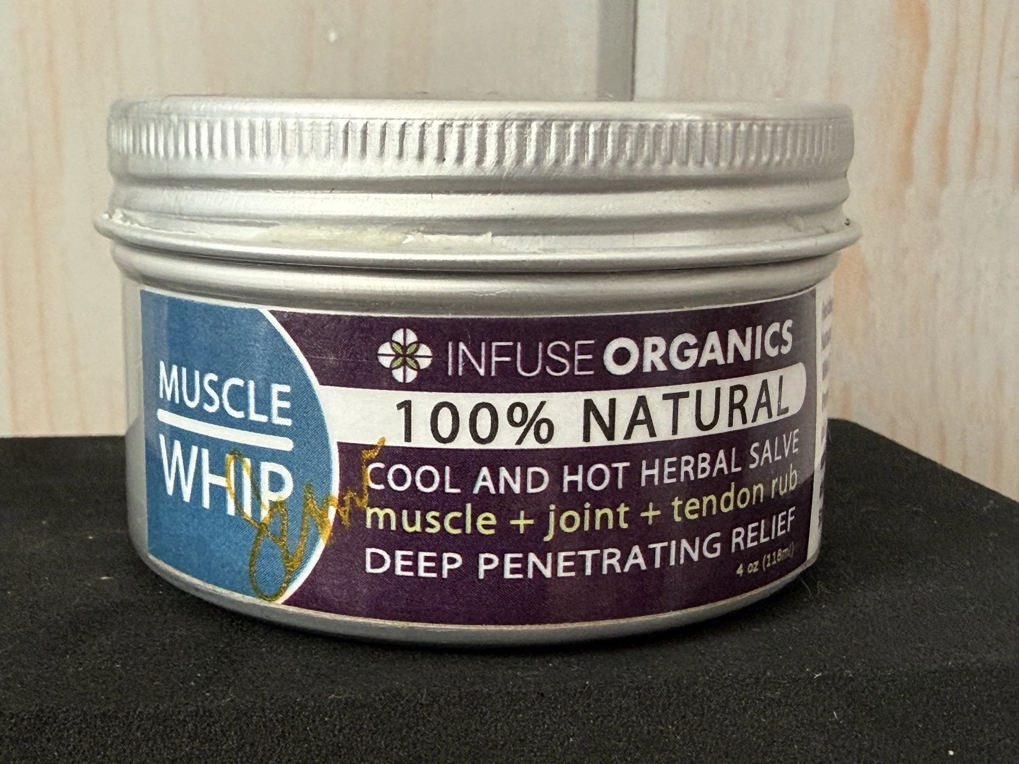 Close-up view of a container labeled 'Muscle Relaxing Whole Body' herbal remedy, showing the silver lid and part of the label with instructions and ingredients, placed on a black surface against a wooden background.