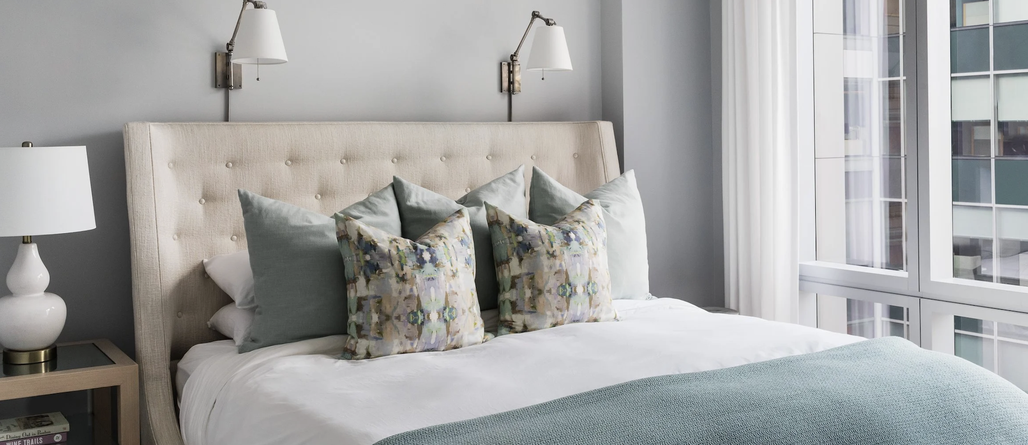 A bright bedroom with a bed dressed in white linens and pillows, quilted throw, and white sconces, and a white lamp on the bedside table.