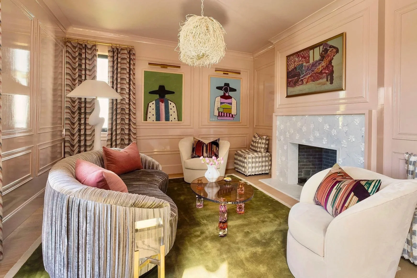 Living room with beige paneled walls, a fireplace, artwork, striped and checkered chairs, a curved sofa with pink pillows, a glass coffee table, and a green carpet.