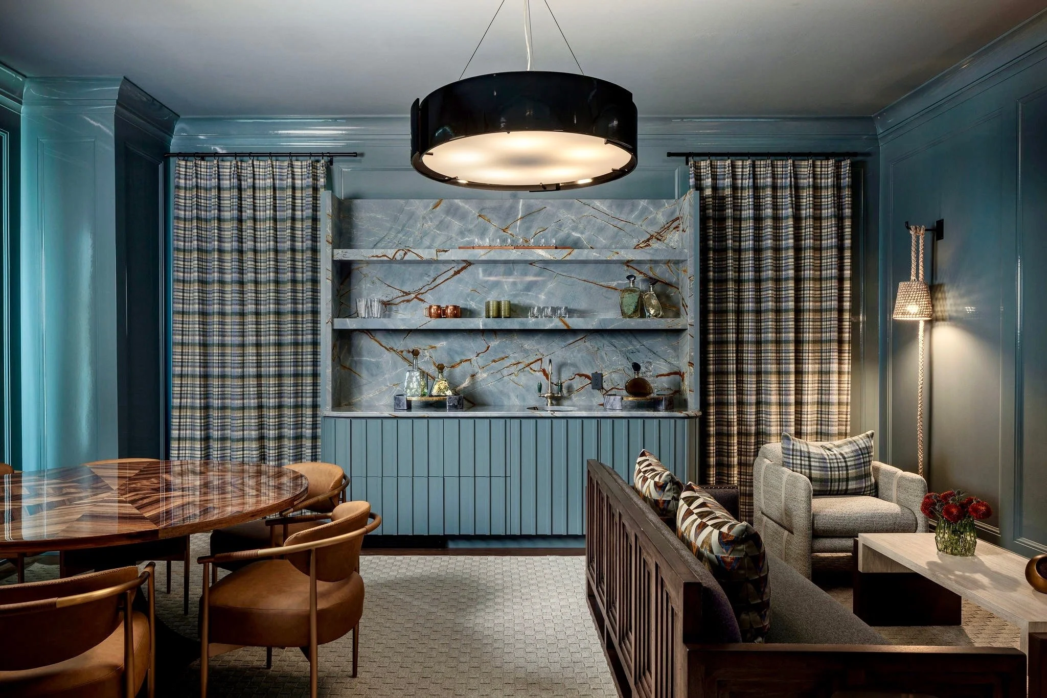 A cozy living room with a marble fireplace, plaid curtains, blue wall paneling, and a round wooden dining table with tan chairs. A black drum-shaped ceiling light hangs above, and a beige armchair with patterned pillows is visible. A floor lamp with 