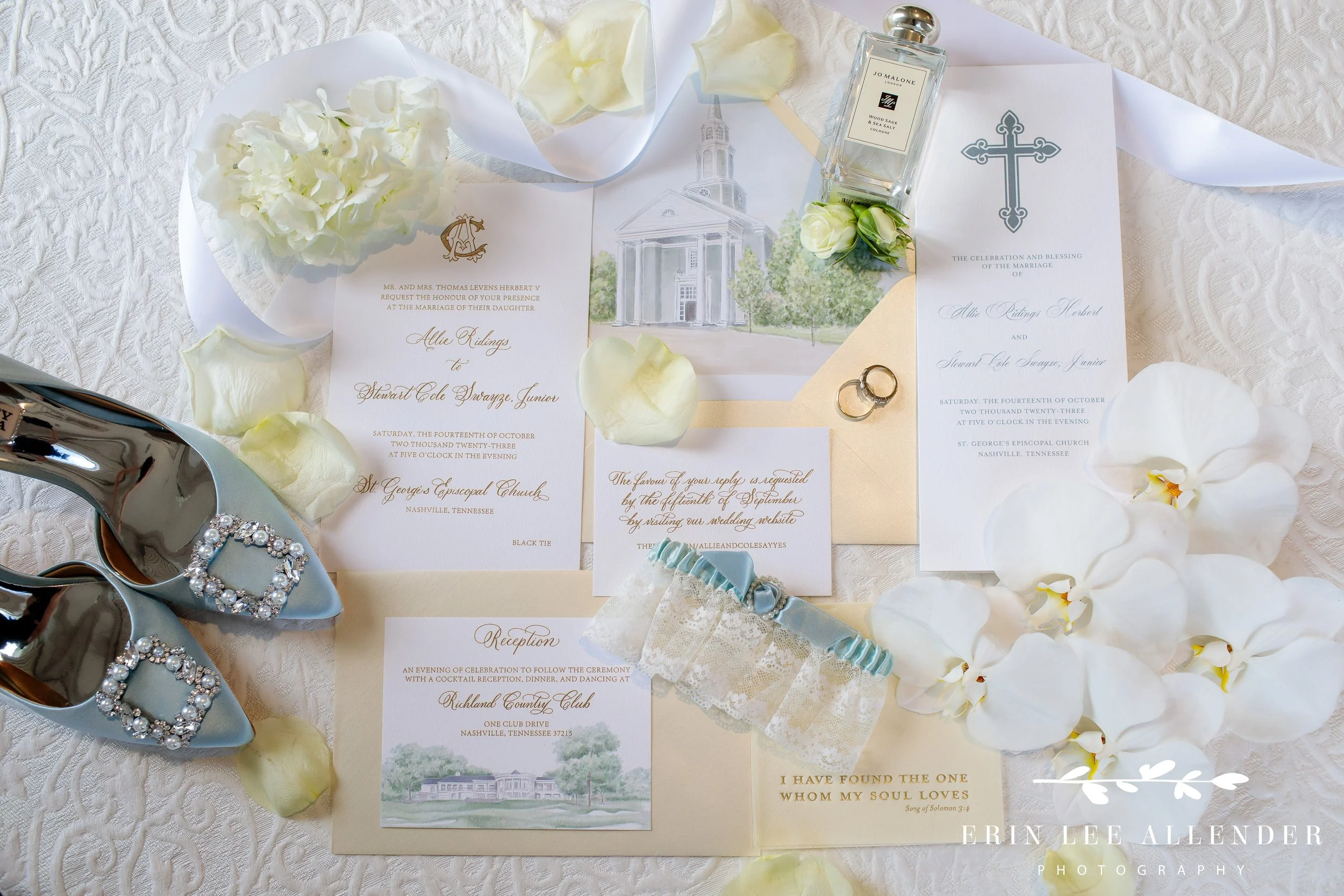 Flat lay of bride’s blue shoes, perfume, and rings on lace robe