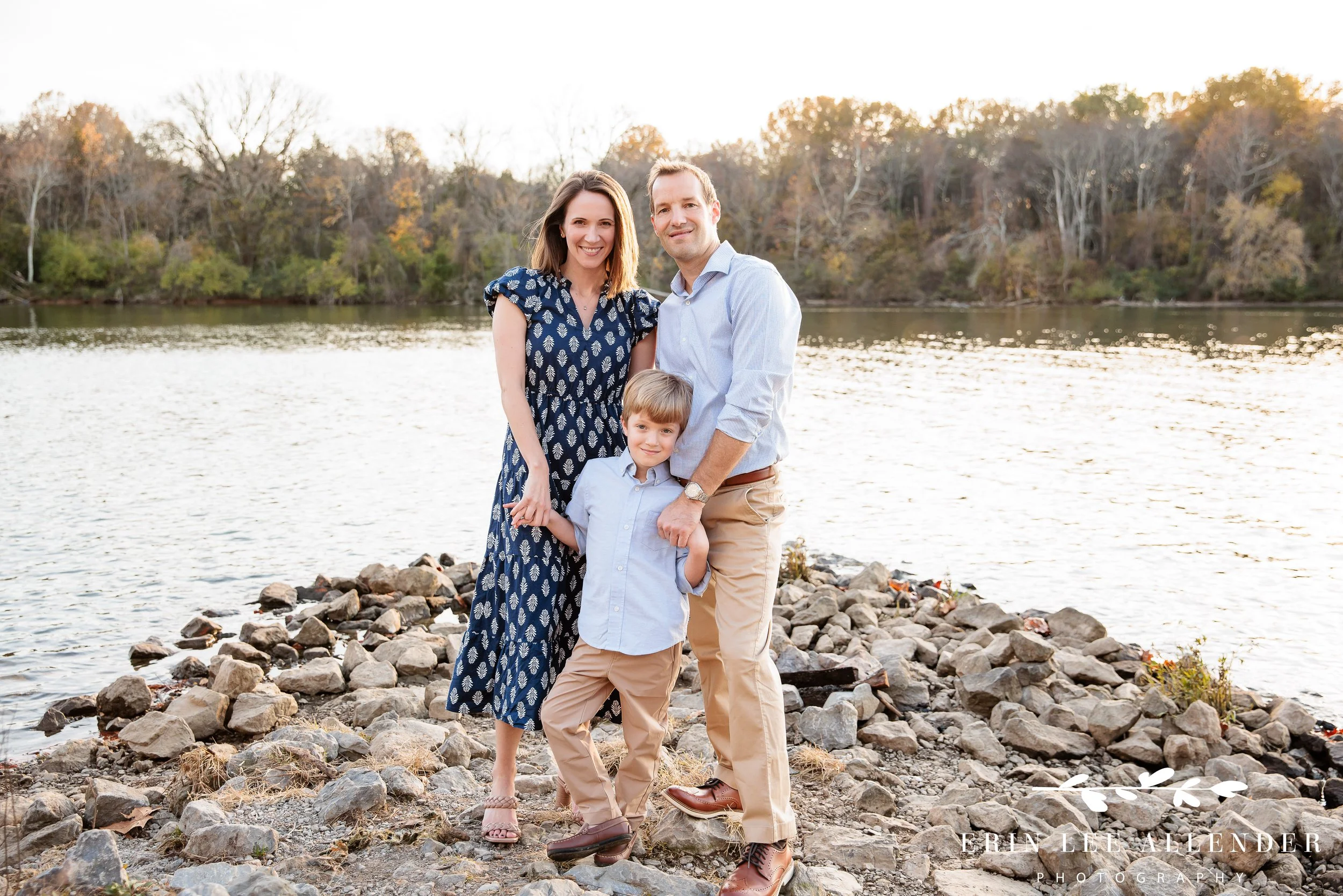 Nashville family photographer's portrait of a family of three standing on rocks with the lake in the background at Walton Ferry Arboretum in late fall