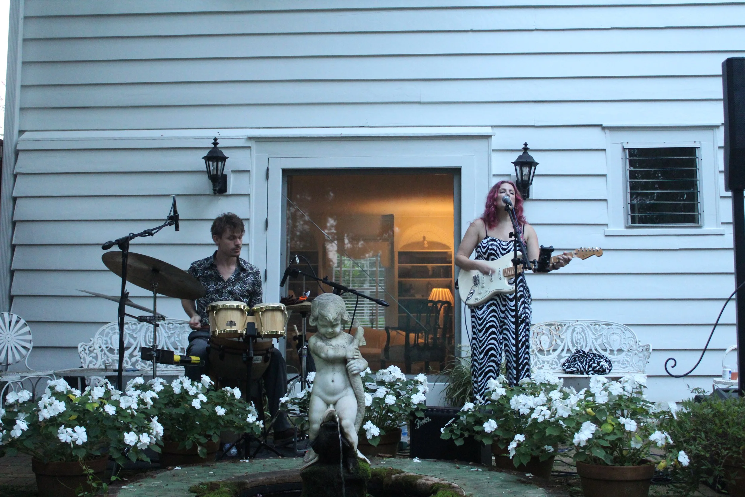 Two musicians playing a guitar and drums playing on a porch behind white flowers and a statue.