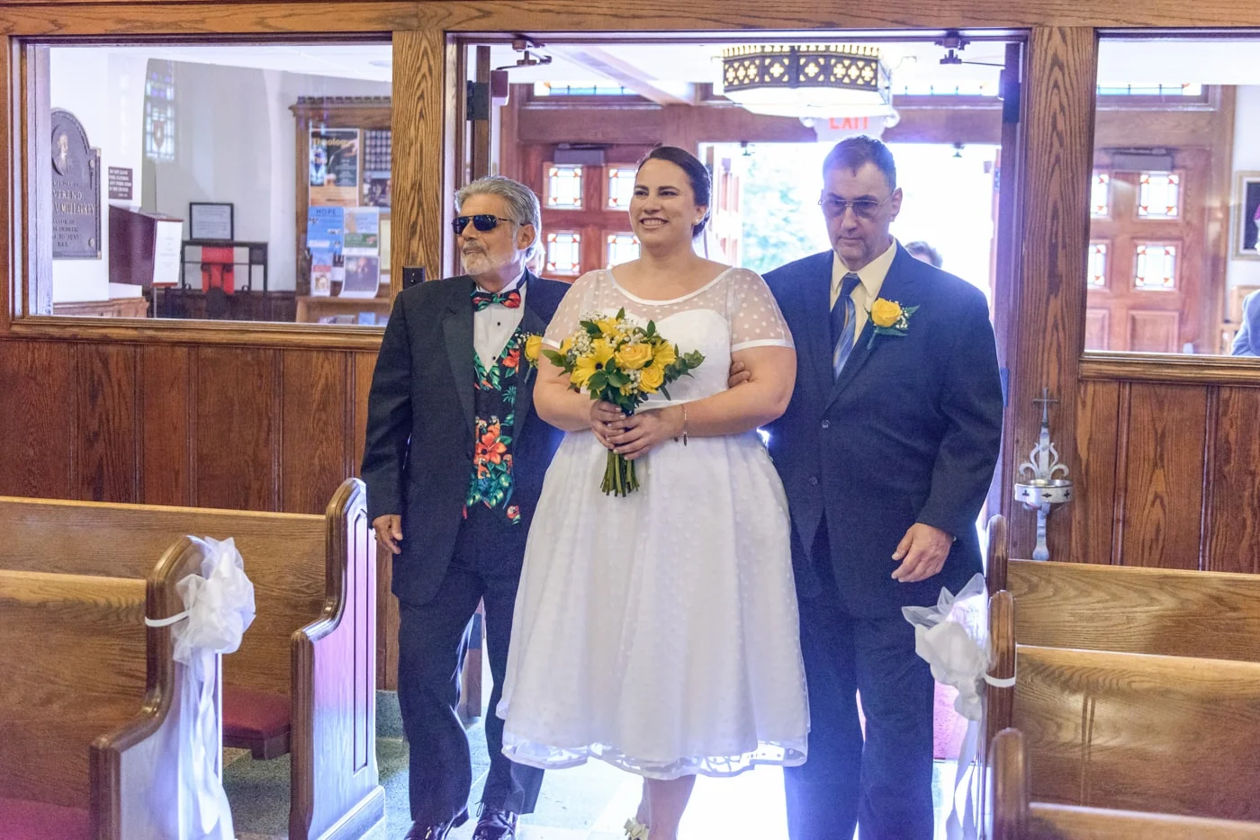 The bride walking in with her father and stepfather holding bright yellow flowers at St James Catholic Church in Falls Church, Virginia