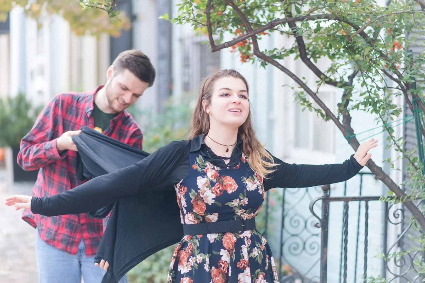 A young woman holds her arms out and looks up like she’s flying as her boyfriend tries to hold her sweater out like it’s flying in the wind in old town alexandria