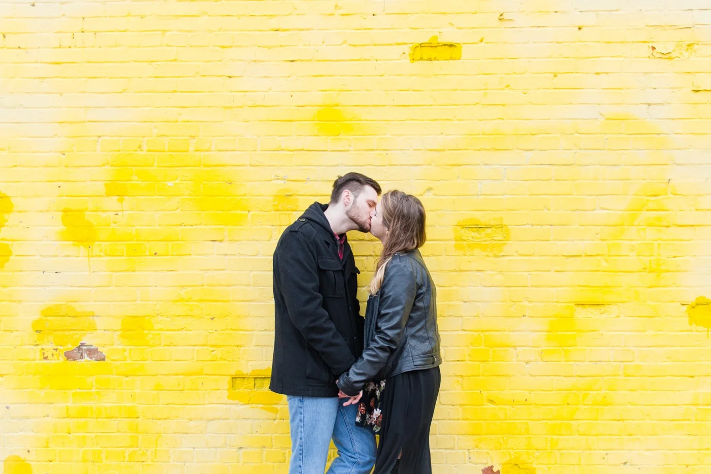 A young couple hold hands and kiss in front of a yellow wall in Old Town Alexandria