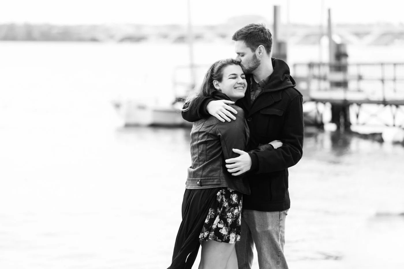 Black and white of a young couple embracing as he kisses the top of her head by the waterfront in Old Town Alexandria