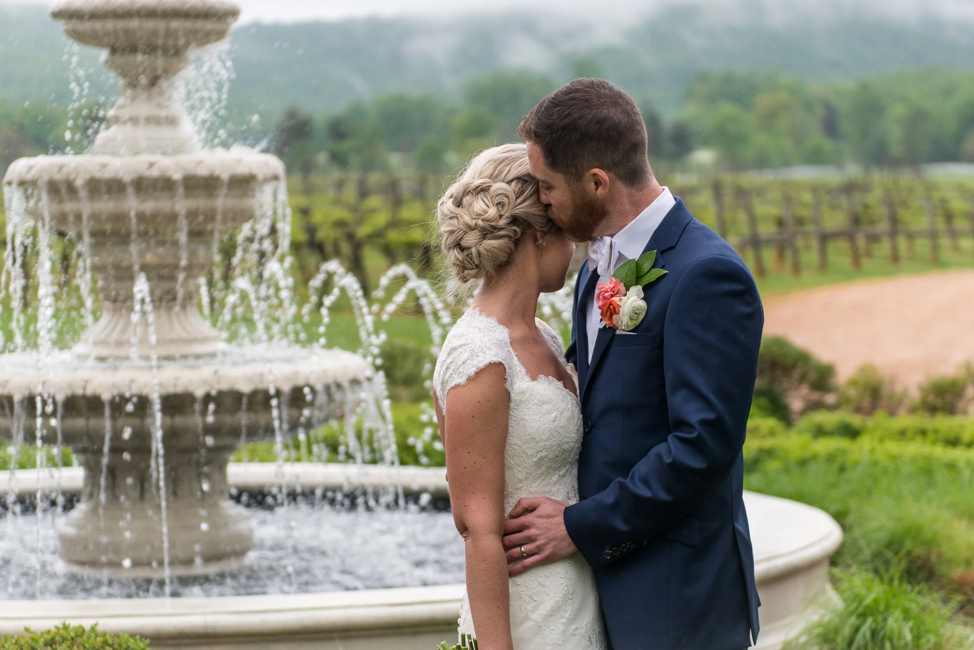 The bride and groom embrace as he kisses her temple next to a water fountain outside Keswick Vineyards in Charlottesville, Virginia