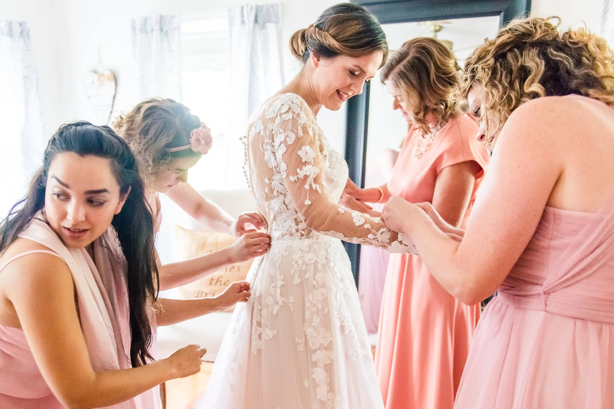 Bridesmaids in pink help the bride put on her modest lace wedding dress in the bridal suite at The Barns at Hamilton Station Vineyard in Leesburg, Northern Virginia before her traditional Catholic wedding