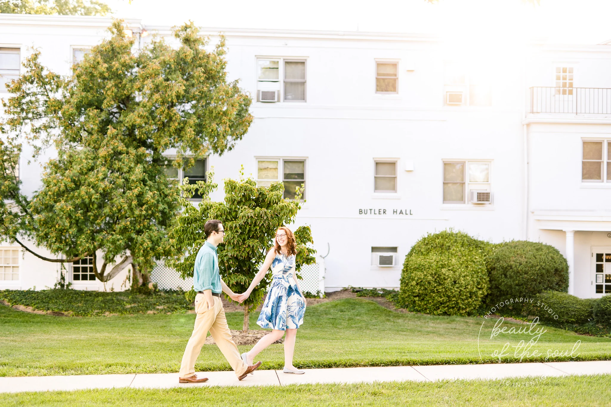 An engaged couple holds hands as they walk in front of Butler Hall at Marymount University during their summer engagement session in Arlington, Northern Virginia