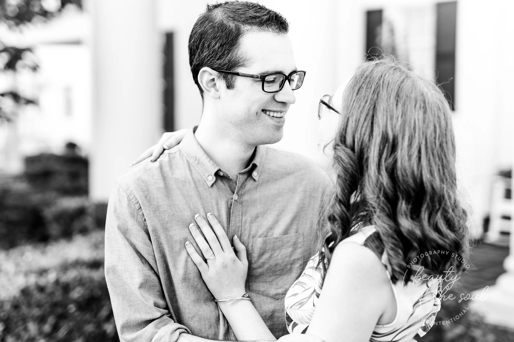 An engaged couple smile and embrace as they stand in front of the Main House at Marymount University during their summer sunrise engagement session in Arlington, Northern Virginia