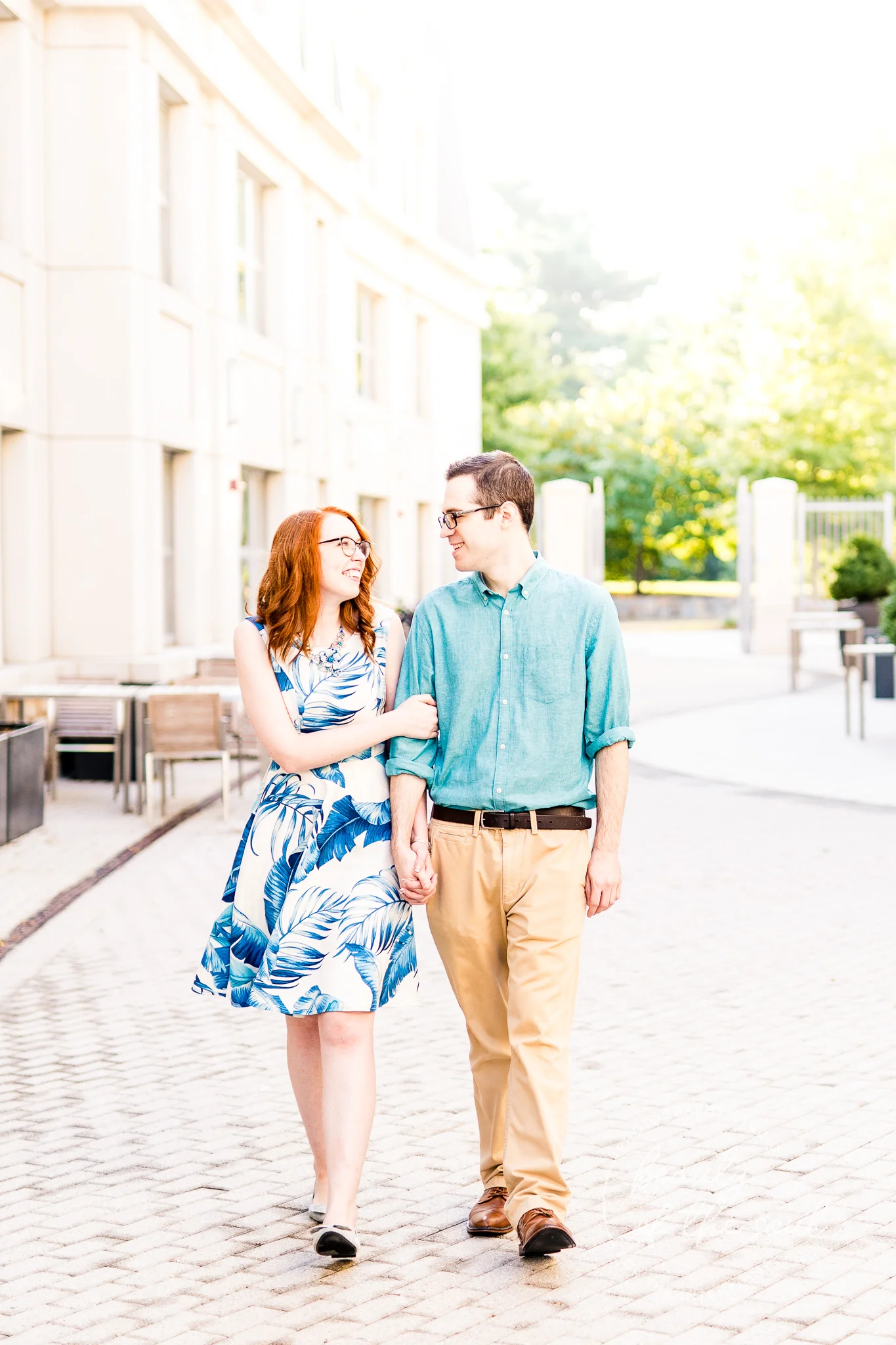 An engaged couple smile at each other as they walk through Malek Plaza at Marymount University during their summer sunrise engagement session in Arlington, Northern Virginia