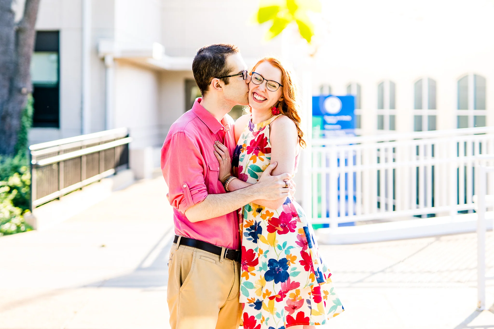 An engaged man kisses the cheek of his smiling fiancee as they stand in front of Gailhac Hall at Marymount University during their summer sunrise engagement session in Arlington, Northern Virginia