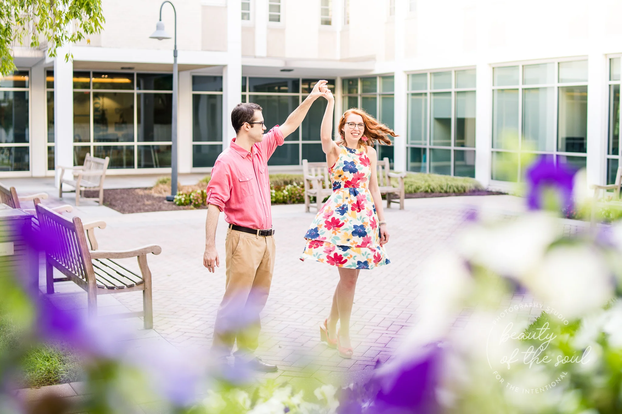 An engaged couple dances together in the courtyard beside Rowley Hall at Marymount University during their summer sunrise engagement session in Arlington, Northern Virginia