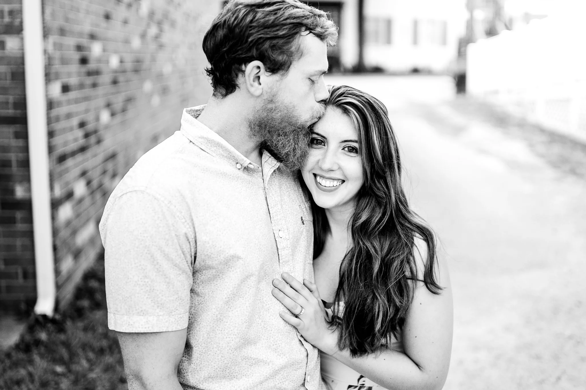 A man kisses a woman on the temple as she smiles during their summer sunset engagement session in Historic Occoquan, Northern Virginia