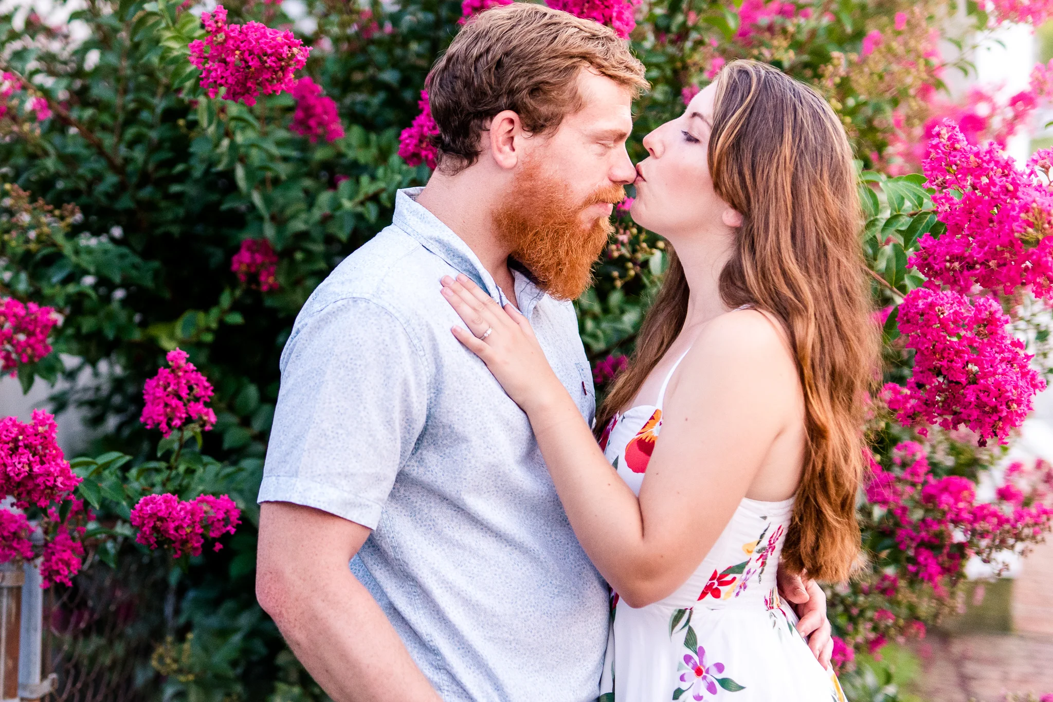 A woman kisses her fiance on the nose as they stand in front of a pink flowering tree during their summer sunset engagement session in Historic Occoquan, Northern Virginia