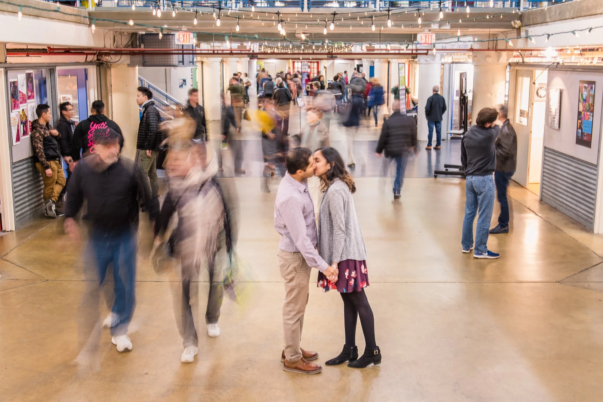 An engaged couple shares a kiss as they stand inside the busy Torpedo Factory Art Center during their engagement session in Old Town Alexandria