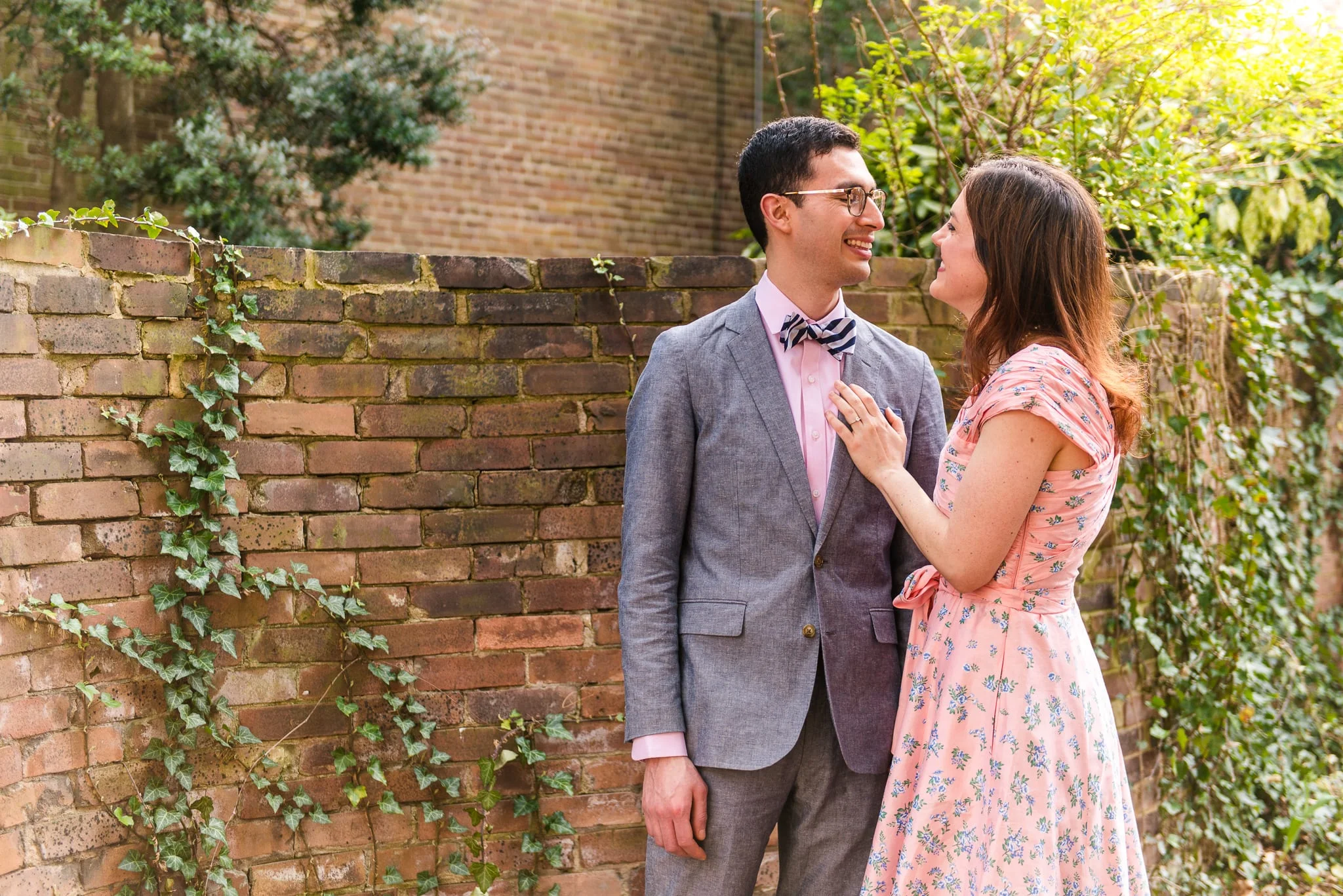 An engaged couple smiles at each other in front of an old brick wall covered in vines during their engagement session in Old Town Alexandria